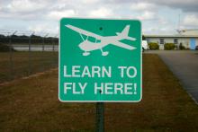 a picture of a green background sign with white lettering. On the sign is an icon of a small plane above the text "learn to fly here"