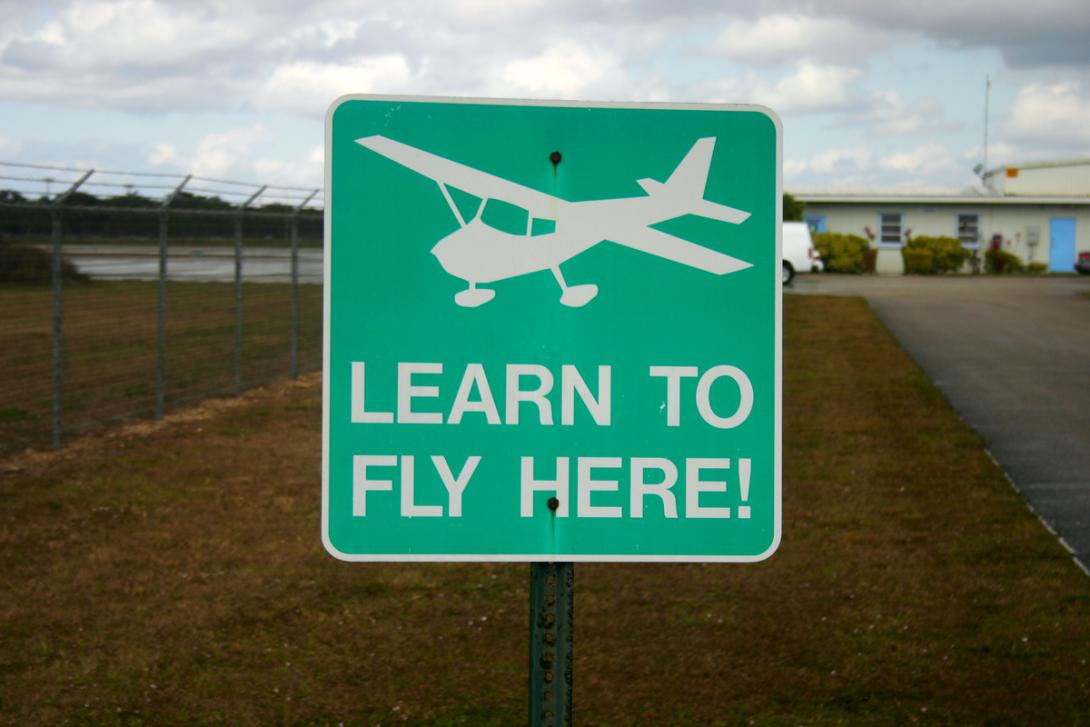 a picture of a green background sign with white lettering. On the sign is an icon of a small plane above the text "learn to fly here"